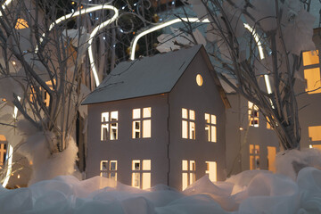 A toy house made of white paper with lights in the Windows. Christmas installation. White snow, lights, garland for the new year.