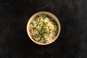 Asian garnish, boiled rice with vegetables, peas, carrots and green onions in a round plate on a black stone table. Pan-Asian and Asian food and dishes on a stylish black concrete kitchen table.