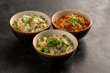 Three Asian side dishes in round bowls of rice, egg noodles, glass noodles and vegetables. Pan-Asian and Asian food and dishes on a stylish black concrete kitchen table.