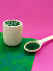Green hawaiian spirulina in tablespoons pills and powder on pink and green background. Super food, healthy lifestyle, healthy supplements concept