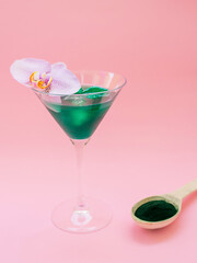 Liquid spirulina green drink with Phalaenopsis in cocktail glass and spirulina pills on pink background. Super food, healthy lifestyle, healthy supplements concept