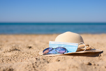 A straw hat, sunglasses and a medical mask lie on the beach by the sea. Focus on the foreground. The concept of a seaside holiday during the coronavirus pandemic.