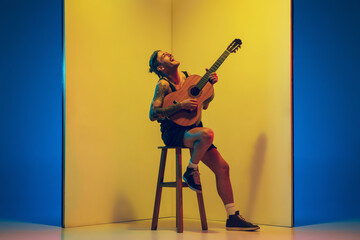 Young musician with guitar performing on yellow background in neon light. Concept of music, hobby,...