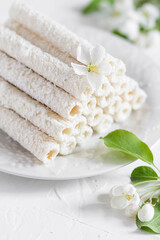 Delicate sweet cookies in the form of tubes in coconut flakes filled with vanilla cream on a white plate. 