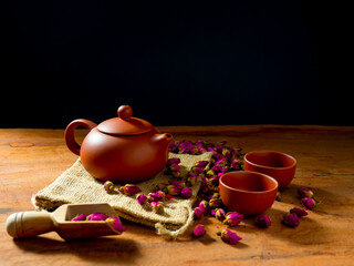 Teapot and teacup with rose tea leaves on wooden table and black background