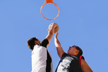 Two young men playing basketball shooting to basket and the other plugging with blue sky background