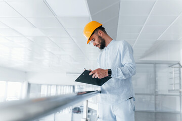 Holding notepad. Engineer in white clothes and orange protective hard hat standing and working indoors