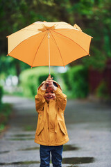 Kid in yellow waterproof cloak, boots and with umbrella playing outdoors after the rain