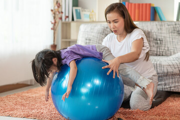 Happy Asian child having fun with fitness ball, doing exercises with mother at home