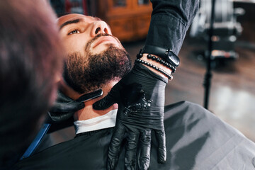 Young man with stylish hairstyle sitting and getting his beard shaved in barber shop