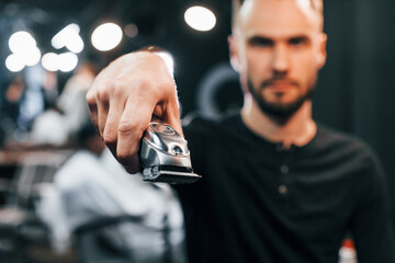 Young bearded man standing in barber shop and holding clipper