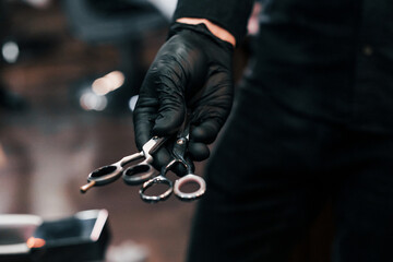 Close up view of guy in black clothes that standing indoors in barber shop and holding scissors