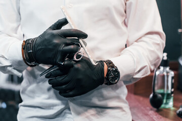 Close up view of guy in white clothes and black gloves that standing indoors in barber shop and holding scissors