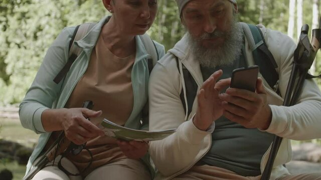 Medium shot of two seniors are sitting in woods with smartphone and map in their hands and discussing path