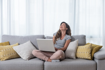 Fototapeta premium Young beautiful single woman chatting with her boyfriend or girlfriend on the laptop computer siting on the sofa and laugh with big smile