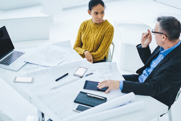 Serious male and female finance professional discussing company situation near desktop with blueprints and smartphone gadgets with blank screen area, entrepreneurs cooperating in modern office