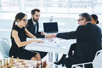 Concentrated team of mature business entrepreneurs communicate near desktop with computer with mock up screen, intelligent architects discussing blueprints buildings for new office of company