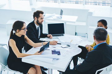 Clever male and female professionals analyzing finance situation during working time in company, team of diversity people collaborating near desktop with modern laptop device with mock up screen