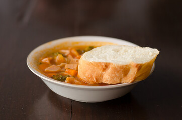 Tomato, red pepper, basil soup in white bowl with bread and spoon on a light gray background.