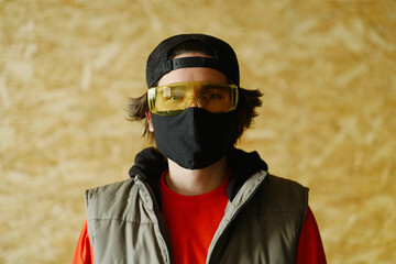 A young man of 25-30 years old in a black protective mask, yellow goggles, a cap and a red jacket posing on a gray background. 