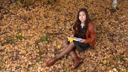 A girl in brown clothes in the autumn forest with a book in her hands. The girl sitting in the park looks up.