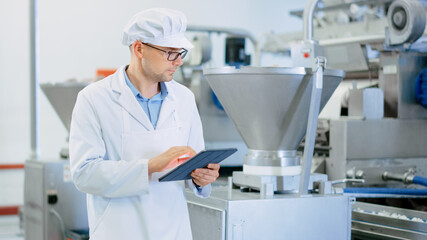 Young Male Quality Supervisor or Food Technician is Inspecting the Automated Production at a Dumpling Food Factory. Employee Uses a Tablet Computer for Work. He Wears White Sanitary Hat and Work Robe. - obrazy, fototapety, plakaty