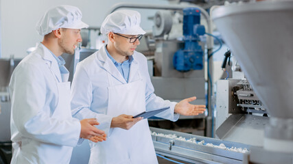 Two Young Male Quality Supervisors or Food Technicians are Inspecting the Automated Production at a Dumpling Food Factory. Employee Uses a Tablet Computer for Work. They Wear White Work Robes.