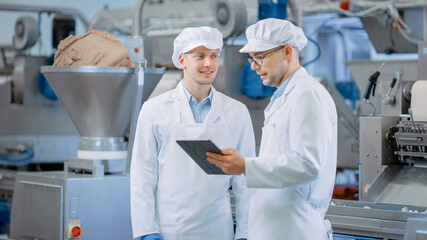 Two Young Food Factory Employees Discuss Work-Related Matters. Male Technician or Quality Manager Uses a Tablet Computer for Work. They Wear White Sanitary Hat and Work Robes.