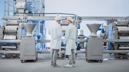 Two Young Food Factory Employees Discuss Work-Related Matters. Male Technician or Quality Manager Uses a Tablet Computer for Work. They Wear White Sanitary Hat and Work Robes.