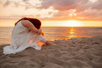 Profile of a single alone or divordes woman silhouette in long white dress sitting on the beach at sunset with her head down.
