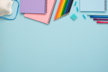 Back to school concept. Colorful stationery set on the blue background. Top view of blue, pink and purple notebooks, paper pins, eraser, felt tip pens, color pencils and face mask. Copy space. 