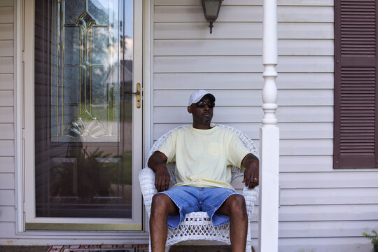 A Portrait Of An  Black African-American Man Sitting Alone In A Wicker Chair On A Porch In Front Of A Home 