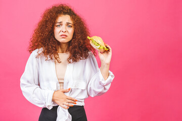Young woman suffer from pain in stomach. She hold bitten burger. Another hand is on stomach area. Isolated on pink background.