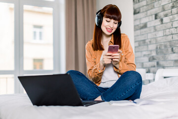 Front horizontal view of dreamy attractive woman with smile texting message on her smartphone, while using headphones and laptop in light stylish room. Working online concept