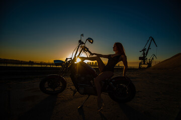 Young redhead woman in erotic lingerie in high heels sits on a motorcycle. Silhouette of a sexy girl on a bike. Industrial landscape.