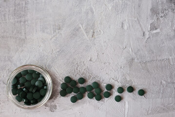 Green chlorella tablets in small glass jar on gray concrete background. Chlorella is a single-celled green algae, it is used to make nutritional supplements and medicine. Top view, copy space.