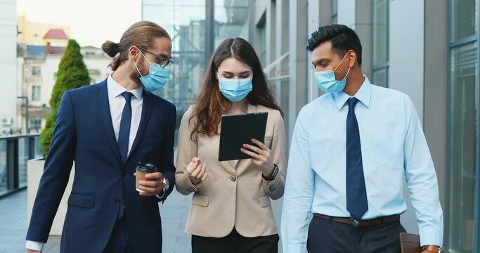 Multi ethnic young businessmen and businesswoman walking the street at business center in medical masks, talking and using gadet. Mixed-races office employees with tablet device strolling outside.