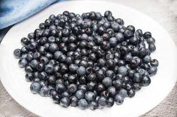 Ripe blueberries lie on a white plate on a light background with a blue napkin. Summer vitamins. Horizontal arrangement. Place for copy space