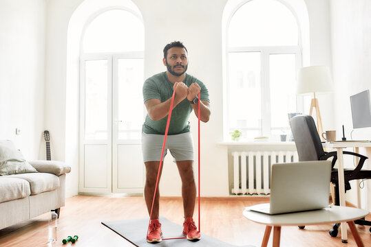 Strive for strong body. Full length shot of male fitness instructor showing exercises with resistance band while streaming, broadcasting video lesson on training at home using laptop