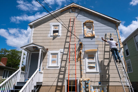 Painter On A Ladder Spraying Paint On The Exterior Of A House