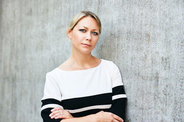 Outdoor portrait of middle age woman, leaning on grey wall, arms crossed
