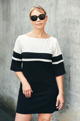 Outdoor portrait of pretty middle age woman wearing black and white dress and sunglasses, posing on grey wall background