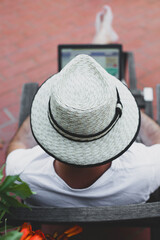 A man in a sun hat sits on the terrace with a laptop.