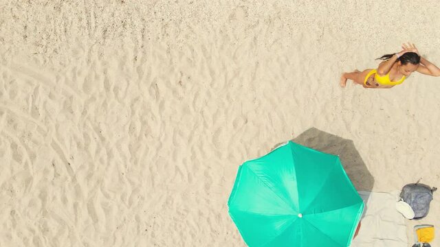 left right top down view drone footage beautiful sexy taned latin woman in bikini standing on white sandy beach and combing her wet hair. clothes, backpack, beach mat and turquoise sun umbrella on