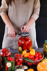 Woman's hands rolls glass preserves with jars sealer. Tomatoes, garlic, pepper, pickles, zucchini, pumpkins on wooden table. Linen apron background. Making homemade vegetables for coronavirus crisis