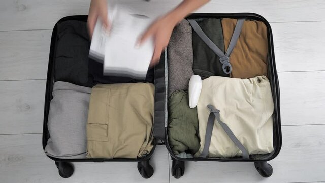 Top view of a suitcase in which women's hands put things for men. Female hands with beige manicure put tablet, camera to pack in a suitcase, the view from the top