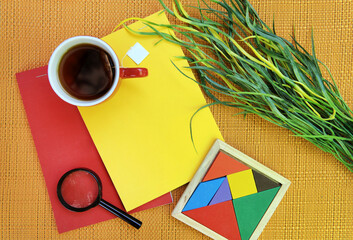 Flat lay top view outdoor photo with bright red and yellow notebooks, tea mug, magnifier and tangram