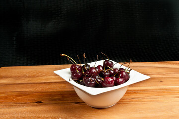 top, front view of a white, deep bowel filled with local, ripe, freshly picked cherries with stems on a wood cutting board and black background