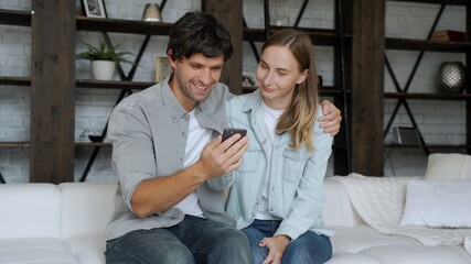 Сouple sitting on sofa at home. Man and woman relaxing on couch with smartphone in living room.