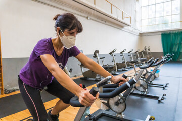 Young caucasian girl with a face mask on a stationary bike in an empty gym due to the low capacity in the new normal, social distance, covi-19, coronavirus